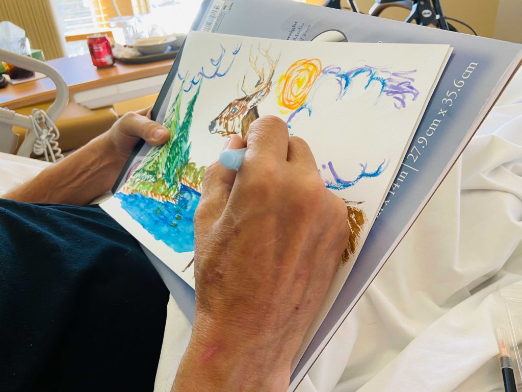 A hospice patient's hands painting a watercolor of a deer, pine trees, and a lake on a sketchpad